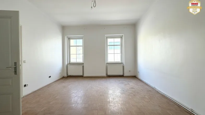 Helles, leeres Zimmer mit Fischgrätparkett und zwei Fenstern, bereit zur individuellen Gestaltung.