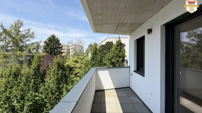 Heller Balkon mit Fliesenboden und Blick ins Grüne, umgeben von Bäumen und Sträuchern.