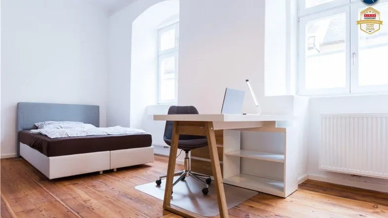Helles Schlafzimmer mit Holzboden, einem Bett und einem Arbeitsbereich am Fenster.