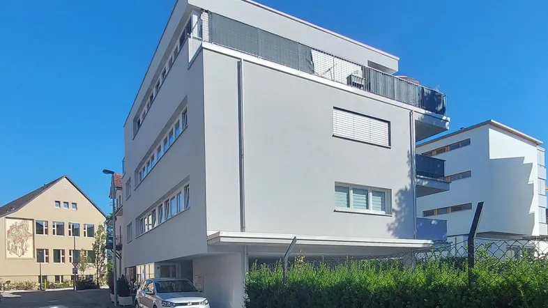 Modernes Mehrfamilienhaus mit heller Fassade, großen Fenstern und überdachten Parkplätzen unter blauem Himmel.