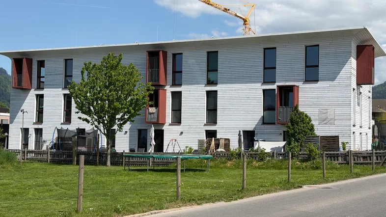 Modernes Mehrfamilienhaus mit weißer Fassade, großen Fenstern und kleinen Balkonen, umgeben von Grünflächen.
