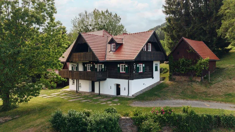 Traditionelles Haus mit rotem Ziegeldach und dunklen Holzfassaden, umgeben von üppiger grüner Landschaft.