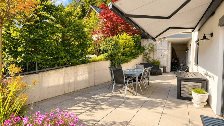 Große gepflasterte Terrasse mit Esstisch, Markise und üppiger Bepflanzung im Hintergrund.