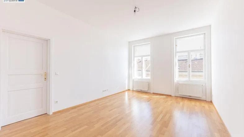 Heller Raum mit Parkettboden, weißen Wänden und zwei großen Fenstern, die viel Tageslicht hereinlassen.