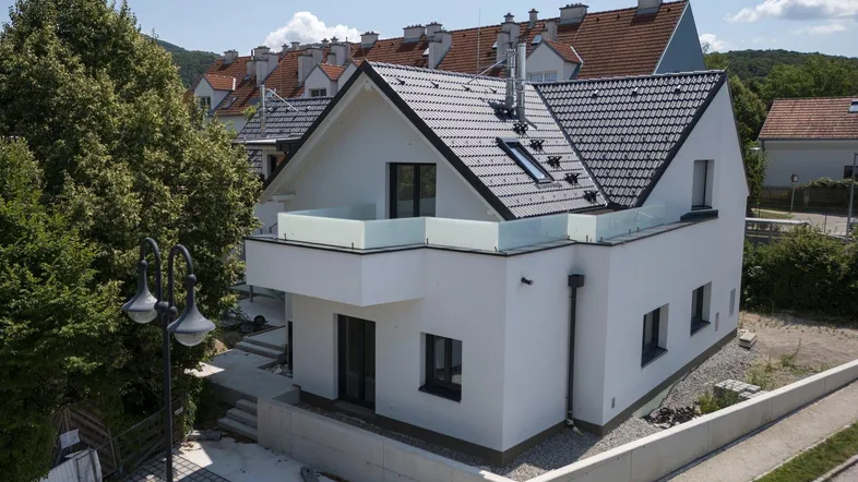 Modernes Einfamilienhaus mit weißer Fassade, dunklem Dach und Balkon in grüner Umgebung.