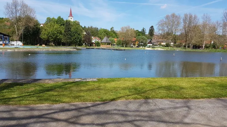 Panoramablick auf einen ruhigen See mit grünen Ufern und einem Dorf im Hintergrund unter blauem Himmel.