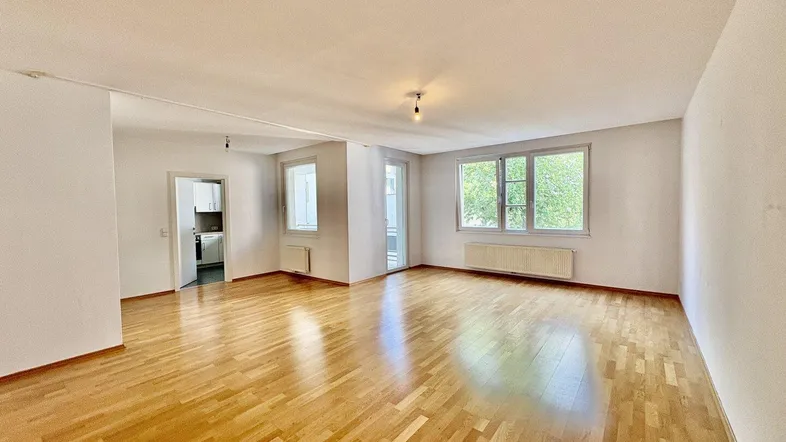 Helles Wohnzimmer mit Parkettboden, großen Fenstern und Zugang zur Küche.