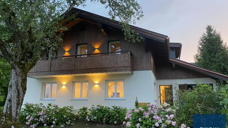 Charmantes Landhaus mit Holzbalkon und blühenden Hortensien im Vorgarten bei Sonnenuntergang.