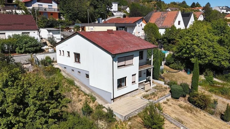 Luftaufnahme eines Einfamilienhauses mit rotem Dach und weißer Fassade in einer grünen Hanglage.