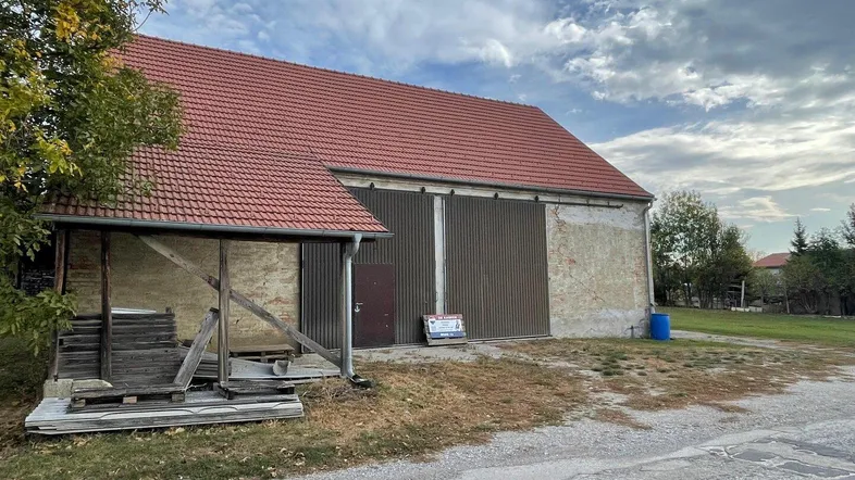 Ländliches Gebäude mit rotem Ziegeldach und großen braunen Toren, umgeben von Vegetation unter bewölktem Himmel.