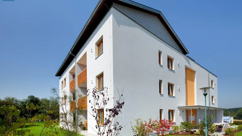 Modernes Mehrfamilienhaus mit weißer Fassade, Satteldach und grüner Gartenanlage unter blauem Himmel.