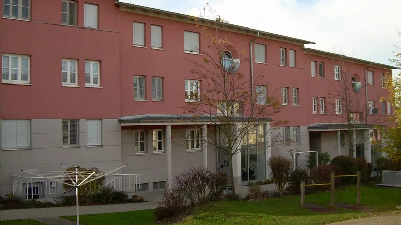 Wohngebäude mit markanter roter Fassade, großen Fenstern und einer gepflegten Rasenfläche im Vordergrund.
