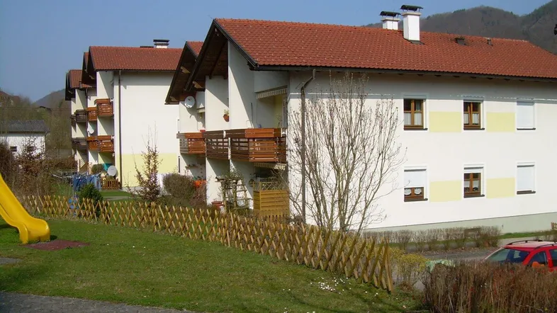 Mehrfamilienhaus mit Satteldach, Balkonen und einem kleinen Spielplatz im Vordergrund, umgeben von grüner Landschaft.