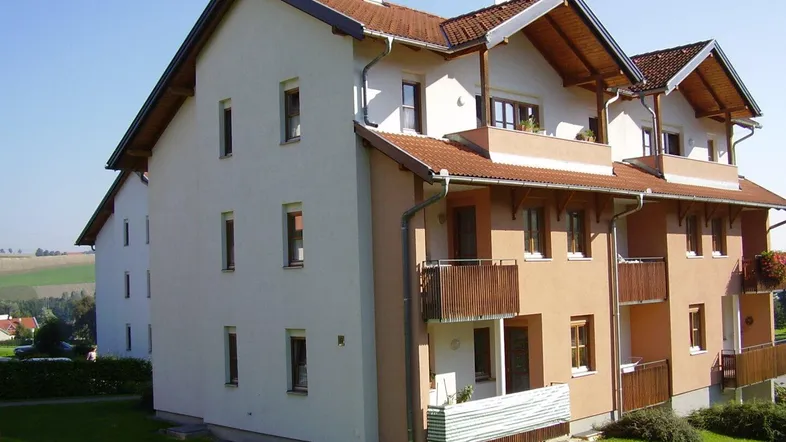Mehrfamilienhaus mit Satteldach, mehreren Balkonen und gepflegtem Rasen, umgeben von ländlicher Landschaft.
