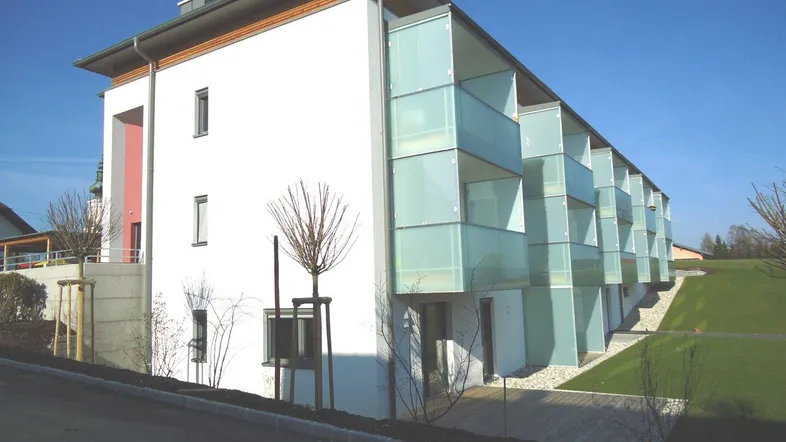 Modernes Mehrfamilienhaus mit weißer Fassade, großen Fenstern und Glasbalkonen in grüner Umgebung.