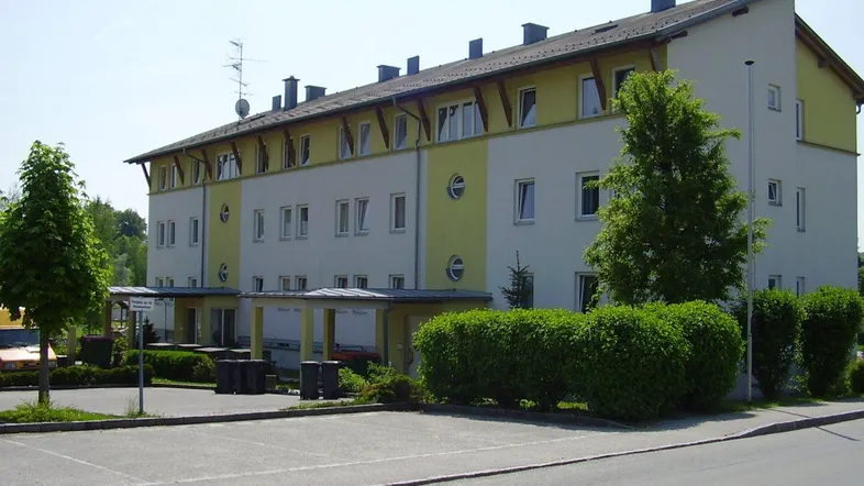 Mehrfamilienhaus mit gelb-weißer Fassade, Satteldach und Parkplätzen im Freien umgeben von Grünflächen.