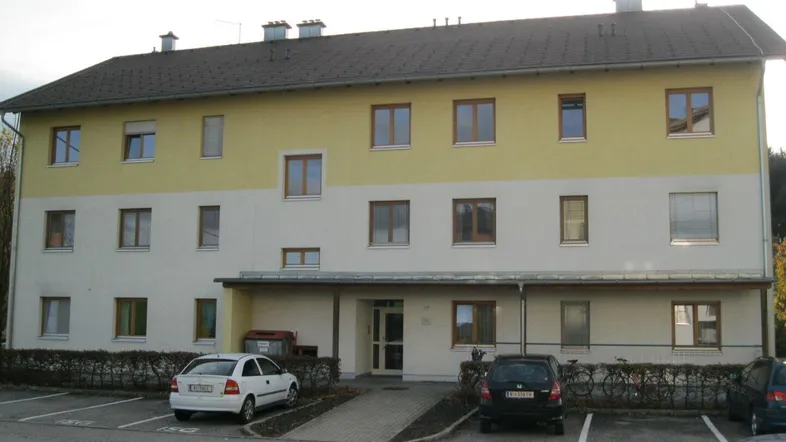 Mehrstöckiges Wohngebäude mit gelb-weißer Fassade, braunen Fenstern und Parkplätzen im Vordergrund.