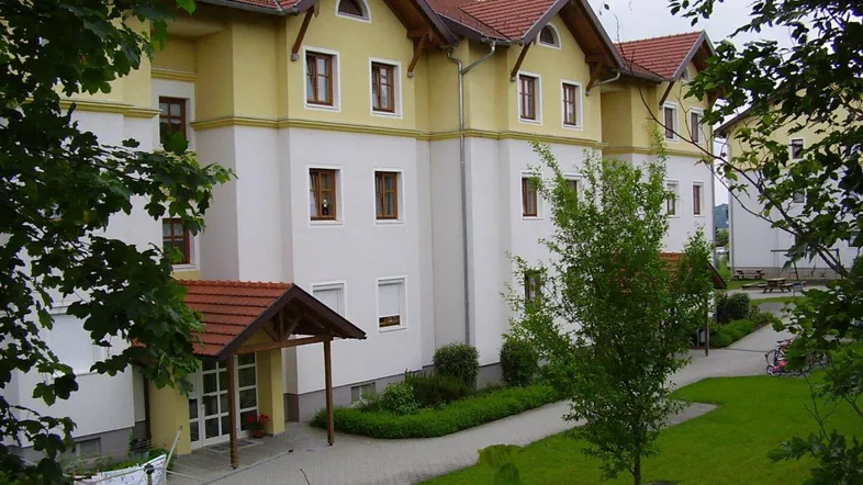 Mehrfamilienhaus mit gelb-weißer Fassade, roten Ziegeldächern und gepflegter Grünanlage umgeben von Bäumen.
