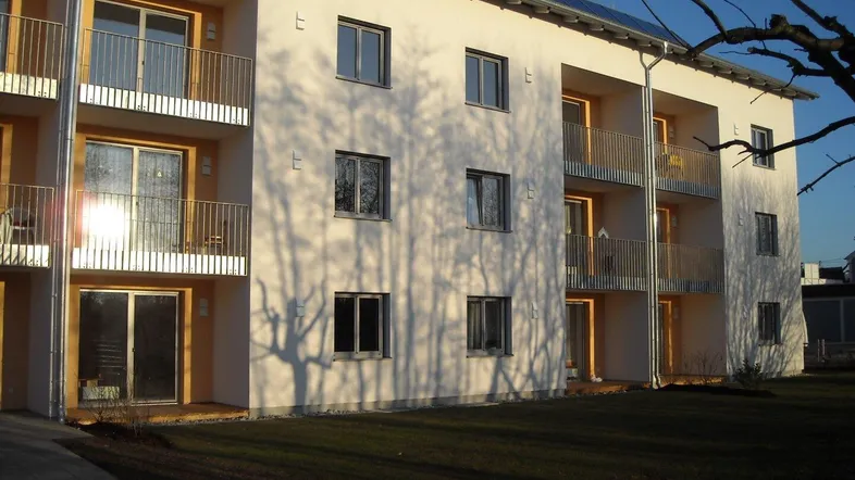 Modernes Mehrfamilienhaus mit Balkonen, großen Fenstern und gepflegter Rasenfläche im Sonnenlicht.
