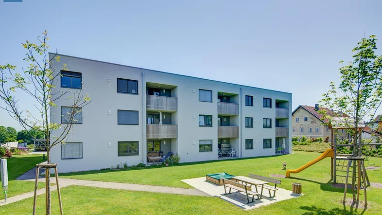 Ansicht eines modernen Wohngebäudes mit Balkonen, großem Rasenbereich und einem Kinderspielplatz mit Rutsche.