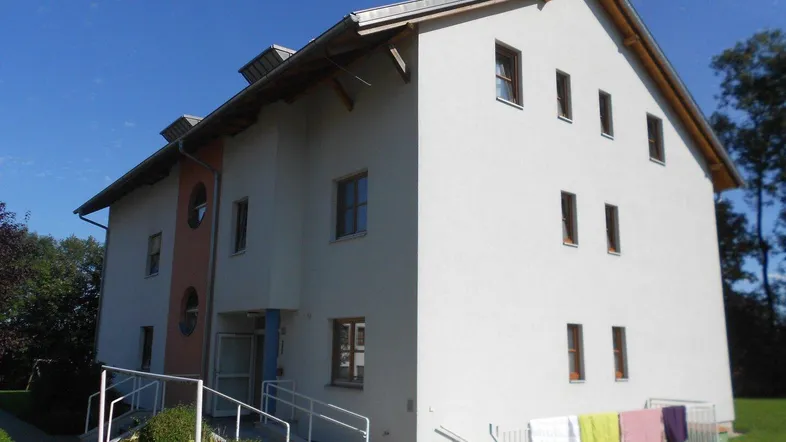 Mehrfamilienhaus mit weißer Fassade, Rampe am Eingang und Satteldach unter blauem Himmel.