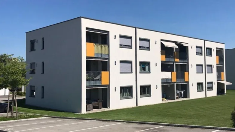 Modernes Mehrfamilienhaus mit Balkonen, großen Fenstern und gepflegter Grünfläche unter blauem Himmel.