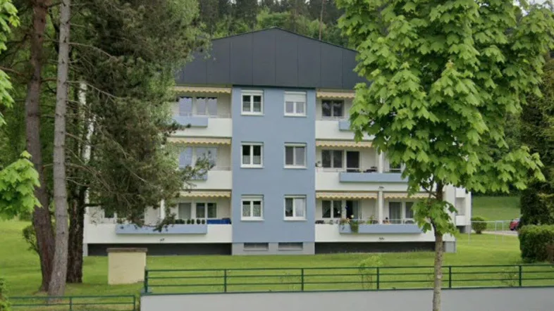 Mehrfamilienhaus mit blauer Fassade, umgeben von viel Grün und Bäumen in ländlicher Umgebung.