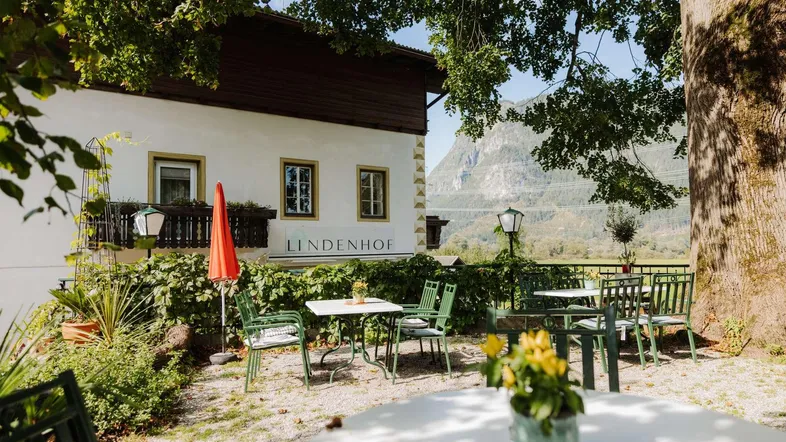 Gemütliche Terrasse mit Gartenmöbeln vor einem traditionellen Gebäude und Bergblick unter Bäumen.