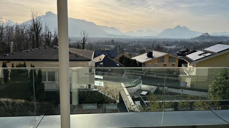 Heller Balkon mit Glasgeländer und weitem Blick über die Stadt und die umliegenden Berge.