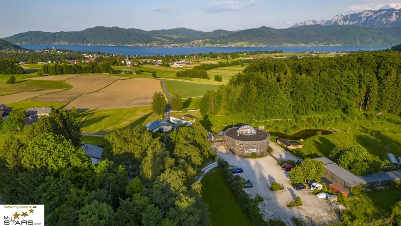 Luftaufnahme des Anwesens mit runden Gebäuden, umgeben von grünen Feldern und einem See im Hintergrund.