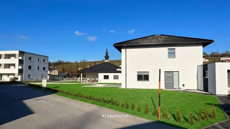 Modernes Einfamilienhaus mit weißer Fassade, schwarzem Dach und gepflegtem Vorgarten unter blauem Himmel.
