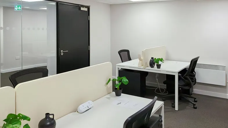 Modernes Büro mit mehreren Arbeitsplätzen, weißen Wänden und Teppichboden, durch eine Glaswand abgetrennt.