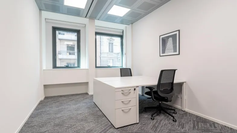 Modernes Einzelbüro mit Teppichboden, zwei Fenstern und einem Schreibtisch mit Bürostuhl.