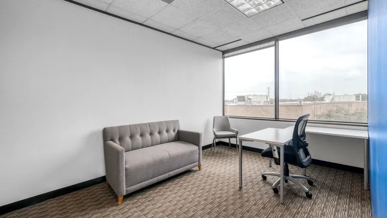 Helles Büro mit Teppichboden, einem bequemen Sofa und einem Schreibtisch am großen Fenster.