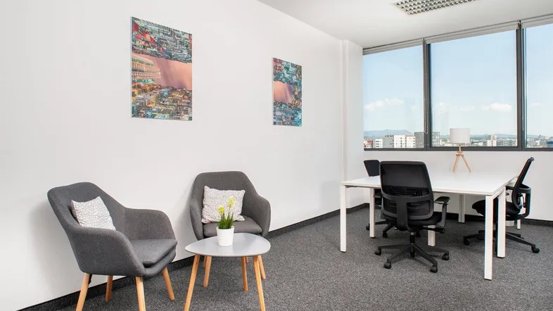 Modernes Büro mit Teppichboden, zwei Sesseln und einem Schreibtischbereich mit Stadtblick.