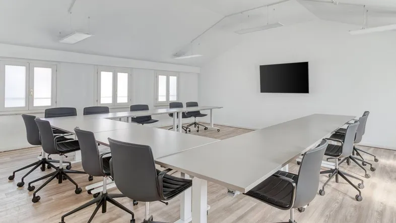 Heller Konferenzraum mit U-förmigem Tisch, modernen Stühlen und einem Wandfernseher für Präsentationen.