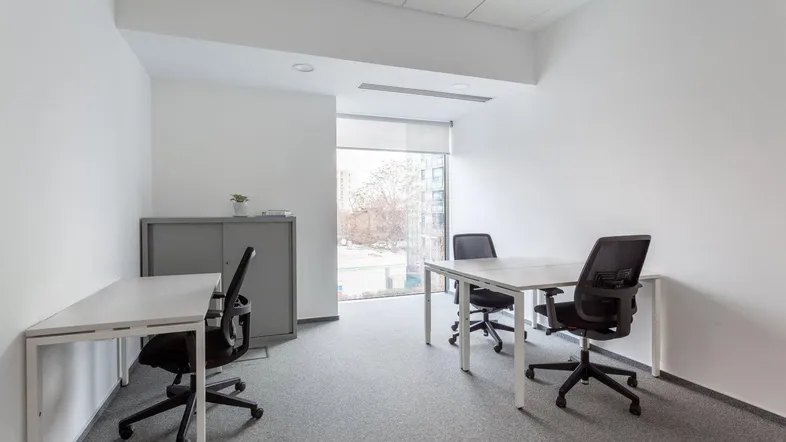 Helles Büro mit zwei Arbeitsplätzen, Teppichboden und einem Fenster, das Tageslicht hereinlässt.