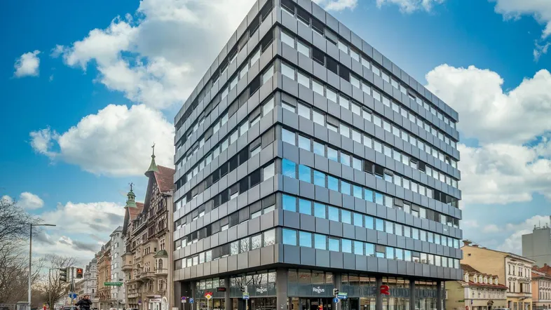 Modernes Bürogebäude mit Glasfassade an einer belebten Straßenecke unter blauem Himmel.
