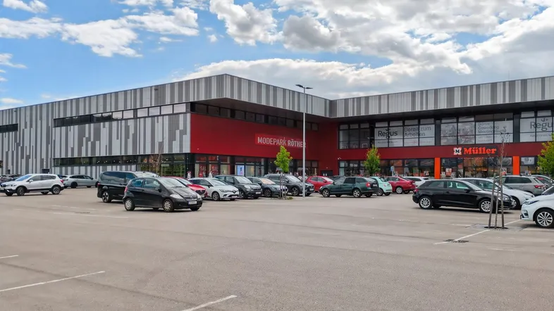 Außenansicht eines modernen Modezentrums mit großem Parkplatz unter bewölktem Himmel.
