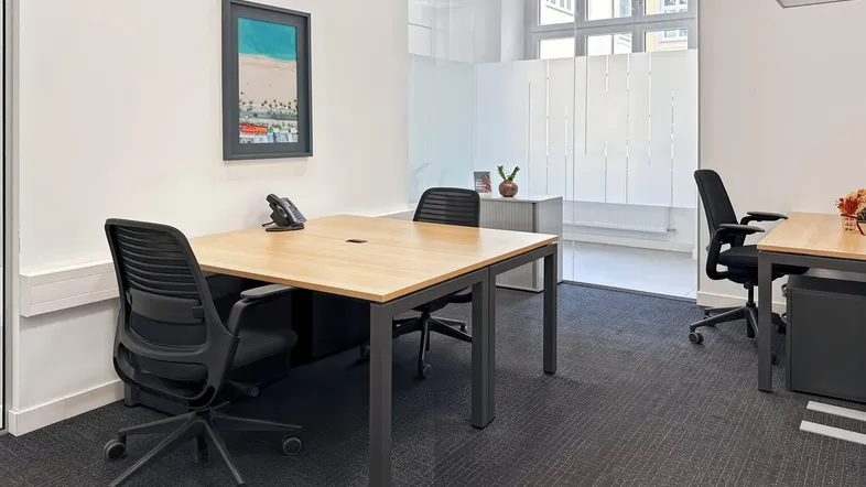 Modernes Büro mit zwei Arbeitsplätzen, Teppichboden und Glaswänden für eine helle und offene Arbeitsumgebung.