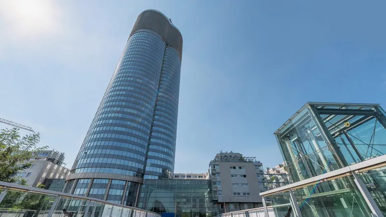 Modernes, zylindrisches Hochhaus mit Glasfassade und einem gläsernen Zugangsweg unter blauem Himmel.