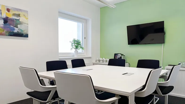 Moderner Konferenzraum mit hellem Tisch, bequemen Stühlen und einem Flachbildschirm an der Wand.