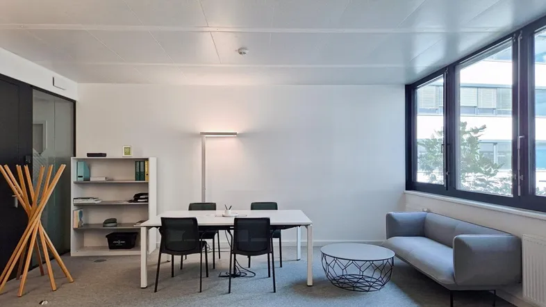Modernes Büro mit Besprechungstisch, Sofa und großen Fenstern, das viel Tageslicht hereinlässt.