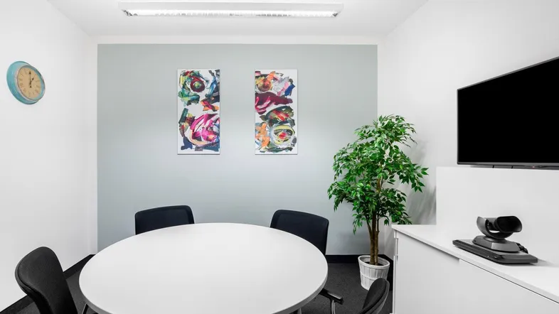 Moderner Besprechungsraum mit rundem Tisch, Videokonferenzsystem und farbenfrohen Kunstwerken an der Wand.