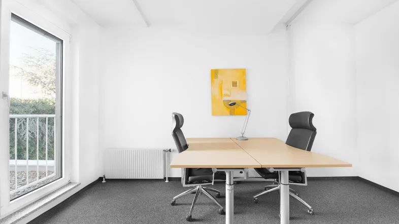 Helles Büro mit zwei Arbeitsplätzen, Teppichboden und einem großen Fenster mit Blick ins Grüne.