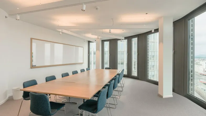 Moderner Konferenzraum mit großem Holztisch, bequemen Stühlen, Whiteboard und weitem Stadtblick durch die Fenster.