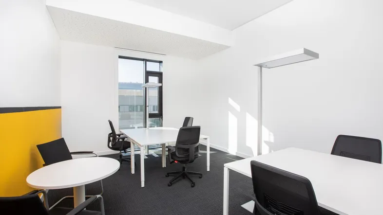 Moderner Büroraum mit weißen Tischen, schwarzen Bürostühlen und einem großen Fenster, das viel Tageslicht hereinlässt.