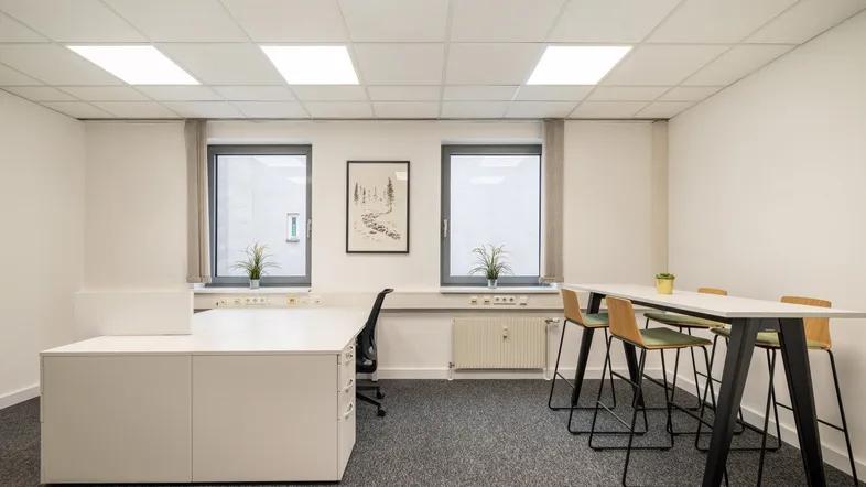 Modernes Büro mit Teppichboden, zwei Fenstern und einer Stehtischgruppe für informelle Besprechungen.