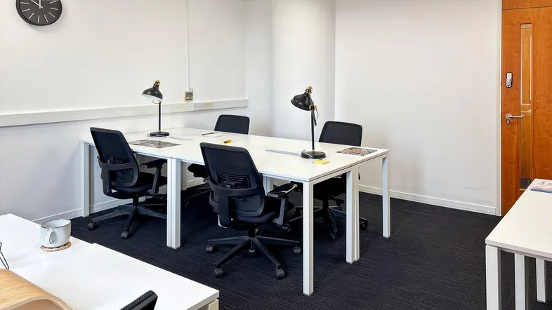 Helles und funktionales Büro mit mehreren Arbeitsplätzen, dunklem Teppichboden und weiß gestrichenen Wänden.