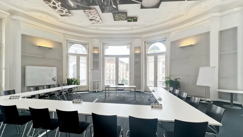 Großer Konferenzraum mit U-förmiger Tischordnung, hohen Fenstern und dekorativer Decke.